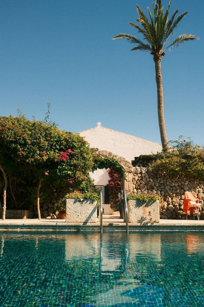 contact-img A serene outdoor pool next to a sunlit stone house with palm tree and lush greenery.